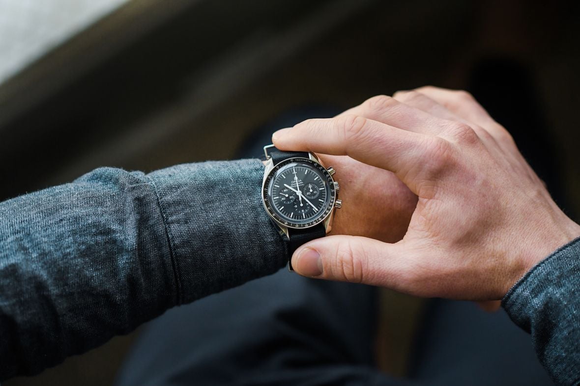 A person's hand wearing a gray jacket sleeve checking the time on an Omega Speedmaster chronograph watch, featuring a black dial, silver case, and textured black leather band.