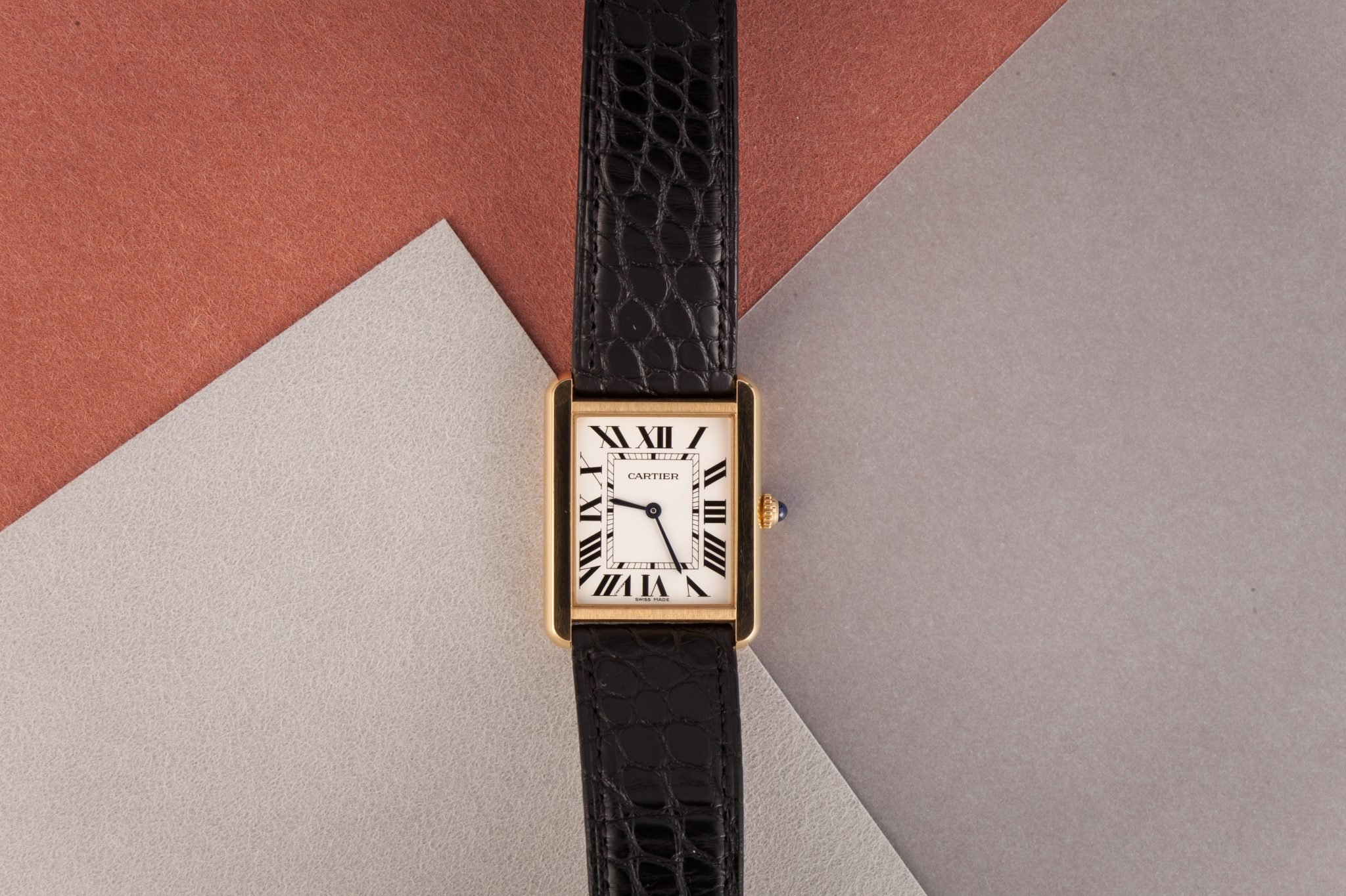 Cartier Tank watch with white square dial and black leather strap on a peach, gray and black background